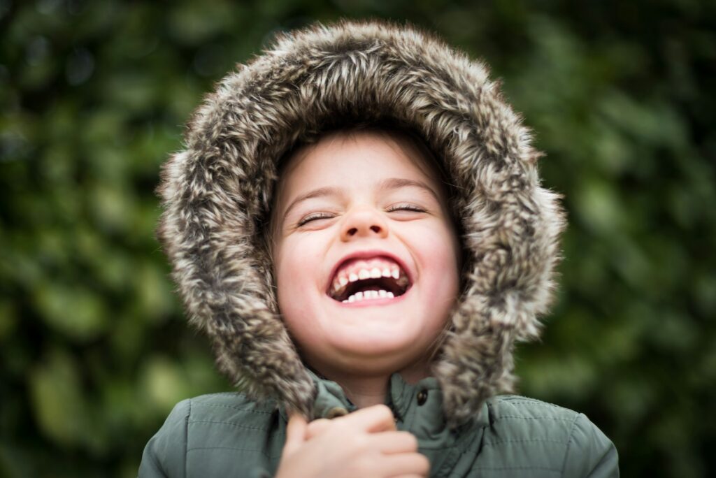 Family Dentist Shenfield Essex. Image of young girl from shoulders up, smiling with eyes closed. She's wearing a green coat with hood up, the hood is trimmed with faux fur.
